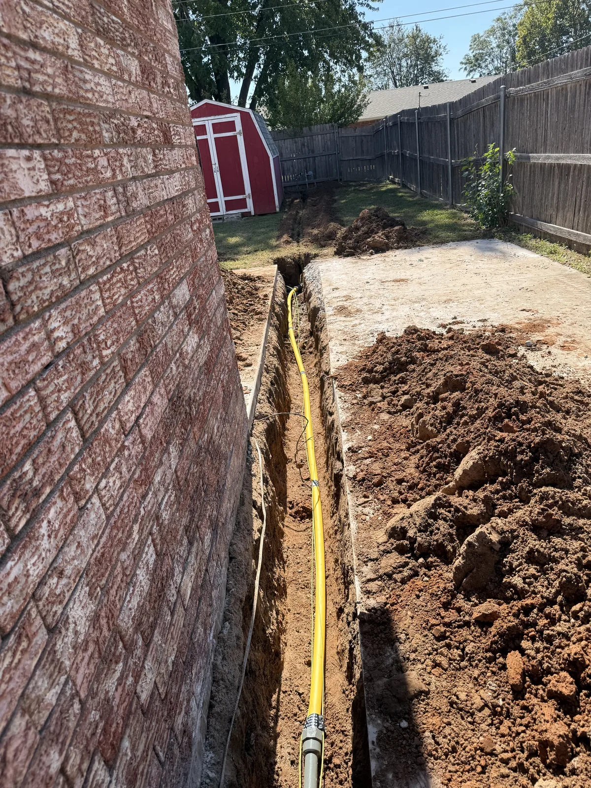 New yellow gas piping in an open trench between the brick house and concrete patio